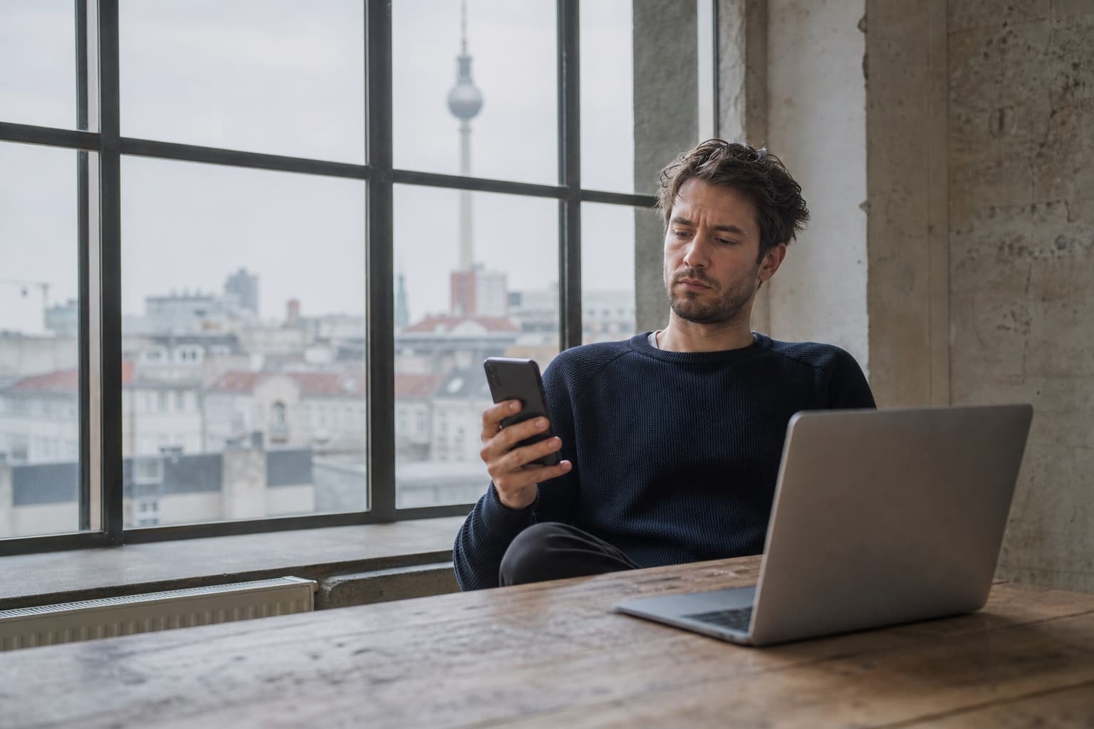 Eine Person sitzt am Laptop, schaut aber abgelenkt aufs Smartphone, im Hintergrund ein Fenster mit Berliner Dächern und Fernsehturm.